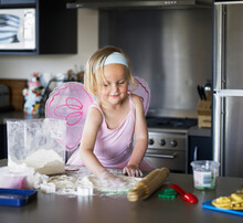 Little Girl Dressed In Ballet Dress And Butterfly Wings Standing At The Kitchen Bench Cooking