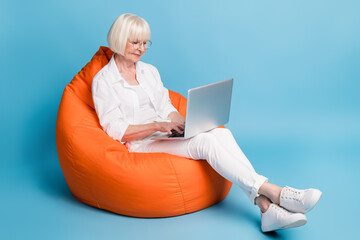 Full length body size photo of senior female entrepreneur in beanbag working on laptop typing isolated on bright blue color background