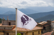 Corsican Flag In Front Of Houses And Mountains In Central Corsica. High Quality Photo
