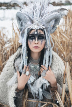 Beautiful Woman With Black Hair And Fantasy Make-up, In Crown With Braids, Horns And Feathers, In White Clothes In Ethnic Boho Style And In Fur Cape Sitting In The Reeds In The Falling Snow