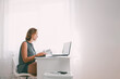 © _KUBE_ - A young woman sitting at a table and leafing through a book. Nearby are a laptop and office supplies. Interior is white color. Copy space