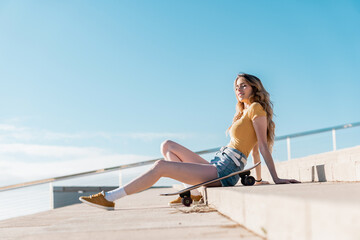 Wall Mural - Long board girl sitting on the stairs outdoors with a skateboard in summer. Sunny day for outside activities. Cool California concept