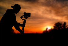 Silhouette Of Videographer Is Filming With Cinema Gimbal Video Dslr At Sunset , Professional Video, Videographer In Events. Cinema Lens On Gimbal. Medium Shot From Right Side. Film Or Cameraman School