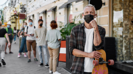 Wall Mural - Portrait of middle aged man wearing mask holding longboard, looking at camera while waiting to collect his takeaway order from the pickup point during lockdown