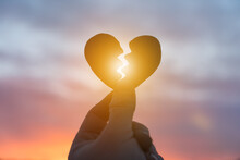 Silhouette Of A Hand Holding A Broken Heart On The Background Of The Sunset. Love Concept, Spat, Divorce.