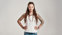 Portrait Of Young Tattooed Caucasian Woman With Dreadlocks Wearing White Shirt, Looking At Camera While Posing Isolated Over Grey Background