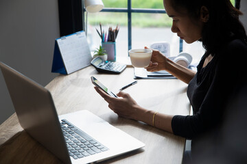 Wall Mural - Remote working at Home office Concept. Businesswoman working online via smartphone and drink coffee. Asian young entrepreneur watching webinars and talkduring meeting video conferences calls with team