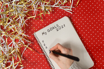 Female hands writing My Goals 2021 in a notebook. Red background and tinsel. New Years concept. Top view