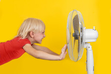 Boy Is Cooling Off With The Fan Wind. Hot Summer Concept. Portrait On Yellow Background