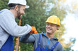 © Friends Stock - Team. Two positive young male engineers in hard hats smiling at each other, giving fist bump while working on cottage construction site outdoors
