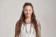 Portrait of young tattooed caucasian woman with dreadlocks wearing white shirt, smiling at camera while posing isolated over grey background