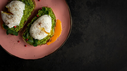 Top view on two poached eggs. Cutted poached egg with liquid yolk on avocado cream on the pink plate. Dark background with copyspace