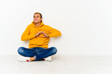 Young caucasian man sitting on the floor smiling and showing a heart shape with hands.