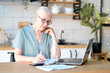 © yavdat - Senior woman using laptop for websurfing in her kitchen. The concept of senior employment, social security. Mature lady sitting at work typing a notebook computer in an home office.