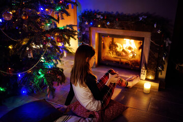 Wall Mural - Happy young girl having a cup of hot chocolate by a fireplace in a cozy dark living room on Christmas eve. Celebrating Xmas at home.