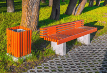 New Orange Bench And Urn In The Sun Near The Sidewalk, Green Grass And Tree Trunks