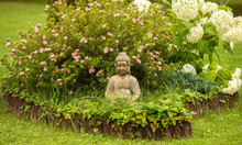 Meditating Buddha Figure Sit Inside Small Island Of Flowers In Flower Bed, Surrounded By Natural Wood Roll Palisade.