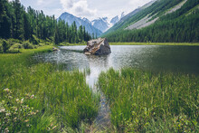 Meditative View To Beautiful Lake With Stone In Valley On Snowy Mountains Background. Scenic Relaxing Green Landscape With Big Mossy Stone In Mountain Lake. Alpine Lake With Ripples On Water Surface.