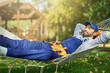 © Friends Stock - Relaxed young male builder wearing blue overalls and cap taking a break, lying in a hammock outdoors with eyes closed on a sunny day