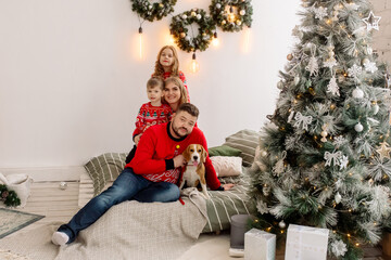 Wall Mural - Young Caucasian beautiful family mom and dad are sitting on the bed with their son, daughter and dog on the windowsill of the house among the Christmas decorations. Family portrait at home
