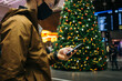 © BublikHaus - Young man in face mask stand in middle of train station, airport or shopping mall decorated with christmas tree. Winter holidays in new normal social distancing. Face mask and smartphone tracing app