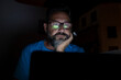 © simona - Stressed and worried man work by night on laptop computer - online social media life and stress for job people - adult male with glasses working with technology at home or office