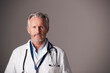 © Monkey Business - Studio Portrait Of Mature Male Doctor Wearing White Coat Standing Against Grey Background