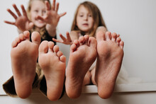 Brother And Sister Holding Bare Feet Close Up To The Camera. Their Blurred Faces In A Background. Hands Reaching To Camera.