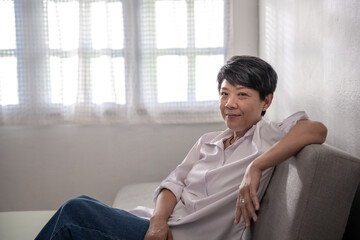 Middle-aged gray-haired Asian woman wearing a white shirt poses at home on a sunny morning, a single retired woman sitting on the sofa in the living room. Positive single senior retired Concept