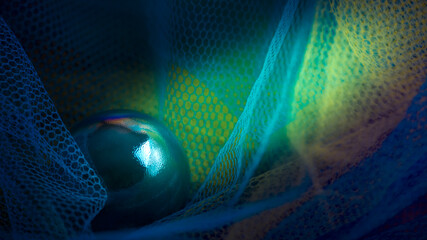 Dark green glass sphere in the darkness of the yellow and blue plastic fishnets. Abstract high resolution macro photography for web design.