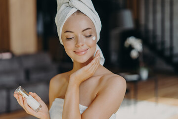 Close up shot of young feminine lady applies face lotion, wears wrapped towel on head, has bare shoulders, smiles with satisfaction, uses cosmetic product. Natural beauty and cosmetology concept