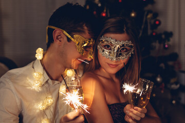 Christmas and New year masquerade party. Couple in love burning sparklers by illuminated Christmas tree with champagne.