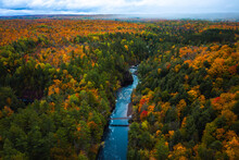 Beautiful Aerial Above The Bad River And A Pedestrian Foot Bridge At Copper Falls With Colorful Fall Foliage Lining The River Banks And Cloudy Sky Above The Horizon In Autumn In Mellen, Wisconsin.