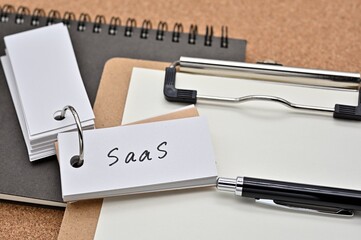 Poster - On top of the notebook and clipboard is a wordbook and pen with the word SaaS written on it. It means Software as a Service.