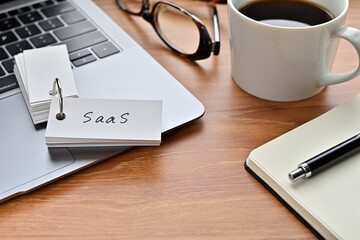 Poster - On a desk with a laptop, glasses, coffee, and a notebook, a vocabulary book was placed open There. The word SaaS is there. It's an acronym that means Software as a Service.