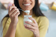 © nenetus - Pretty young woman looking at camera while eating yogurt sitting on the kitchen at home.