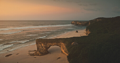 Romantic sun rise at dark rock of Bawana sand beach, sea gulf coastline. Relax seascape sunrise at wavy ocean cliff coast of Sumba Island, Indonesia aerial view. Cinematic soft light drone shot