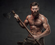Shirtless medieval warlike viking with beard and muscular build posing holding two handed axe in dark studio background.