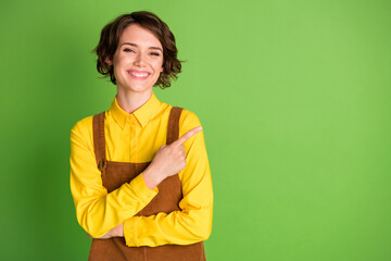 Photo of cheerful girl toothy smile direct finger empty space wear yellow shirt overall isolated green color background