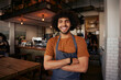 Handsome young male waiter wearing apron standing with crossed arms in cafe looking at camera