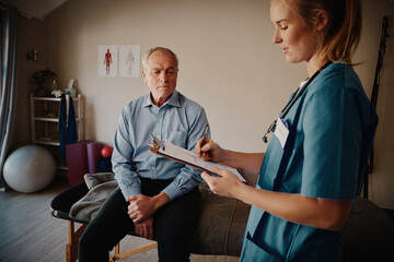 Young woman physiotherapist with stethoscope writing treatment progress of senior male patient on clipboard