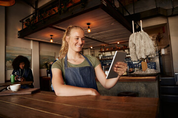 Wall Mural - Smiling female waiter standing leaning on table using digital tablet in modern cafe