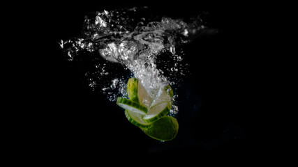 Wedges of lime and slices of cucumber splash into a tank of water on a black background.