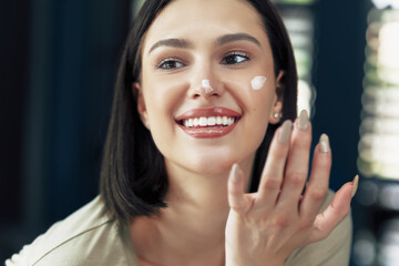 Canvas Print - Happy girl with natural makeup applying cream on her face. Brunette young woman using moisturizing cream.