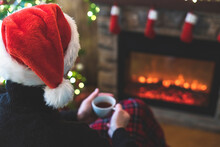 Man With Cup Coffee, Tea In Santa Claus Hat Sitting And Warming At Winter Evening Near Fireplace Flame, Covered Christmas Plaid