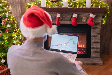 Man freelancer in santa claus hat working on laptop with graphs and charts on screen sitting near christmas tree and fireplace.