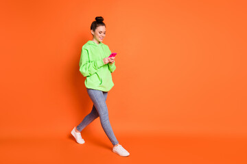 Full length photo portrait of woman texting holding phone in two hands walking isolated on vivid orange colored background