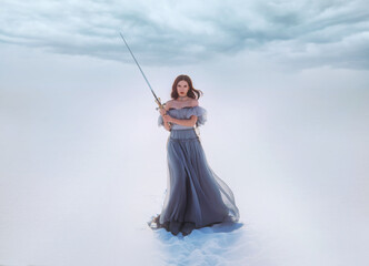 Fantasy woman princess warrior holding medieval iron sword in hands. Fairy tale snow queen. vintage gray blue dress. White winter nature background, valley, tree. Elf girl with steel blade. Art photo.