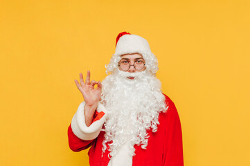 Portrait of a bearded Santa Claus showing an ok gesture and looking at the camera. Santa is expressing an ok symbol with his hand, standing on a yellow background.