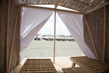 Little Resting Hut Interior With White Curtain And Wooden Benches At Sandy Beach.